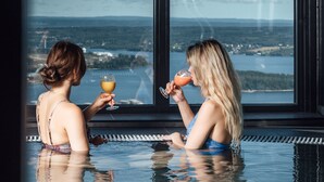 Dos personas en una piscina con fondo de cristal y una vista panorámica de un lago y montañas.