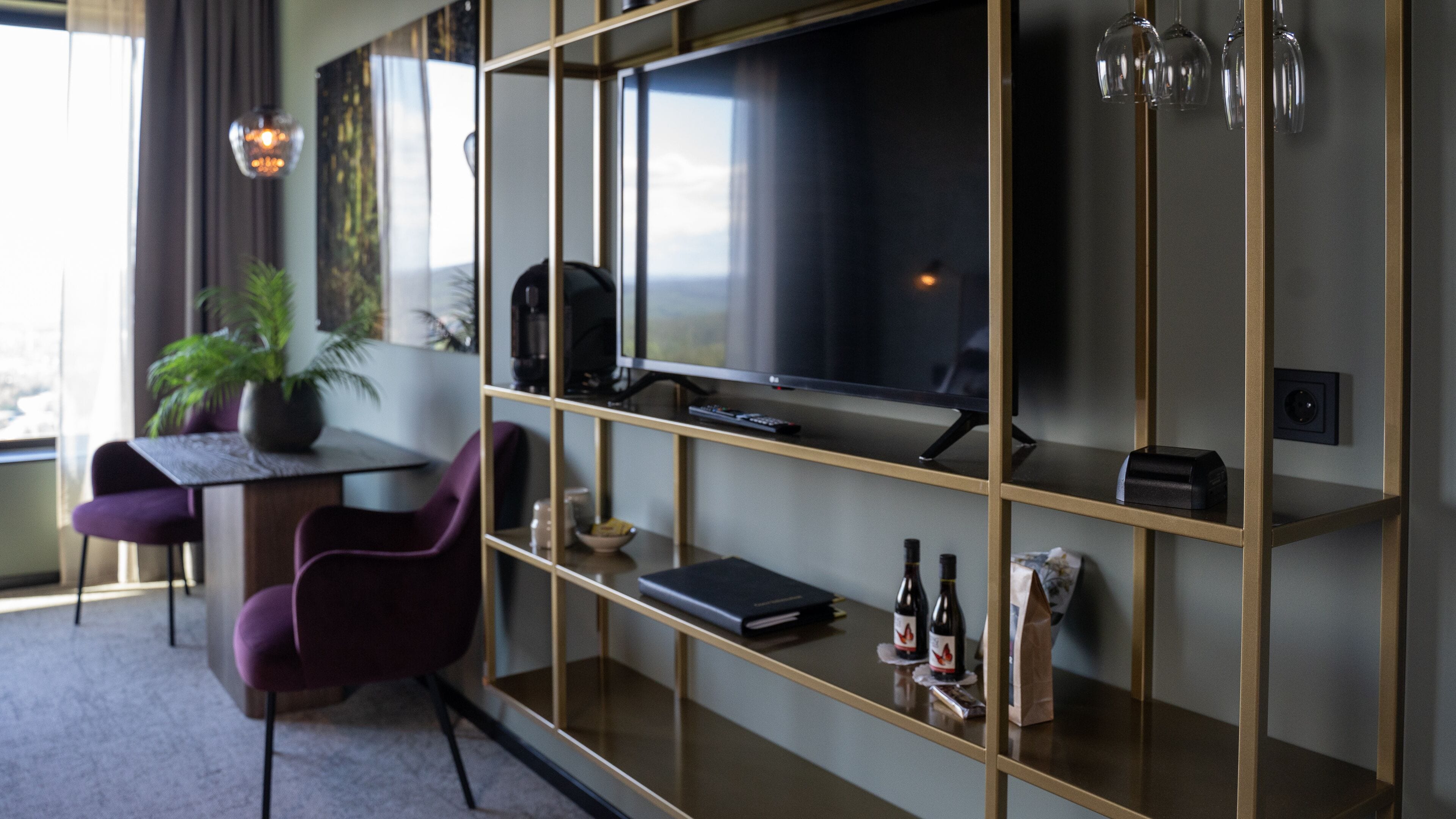 A modern hotel room with a large flat-screen TV, a desk with a chair, a bookshelf, and a potted plant.