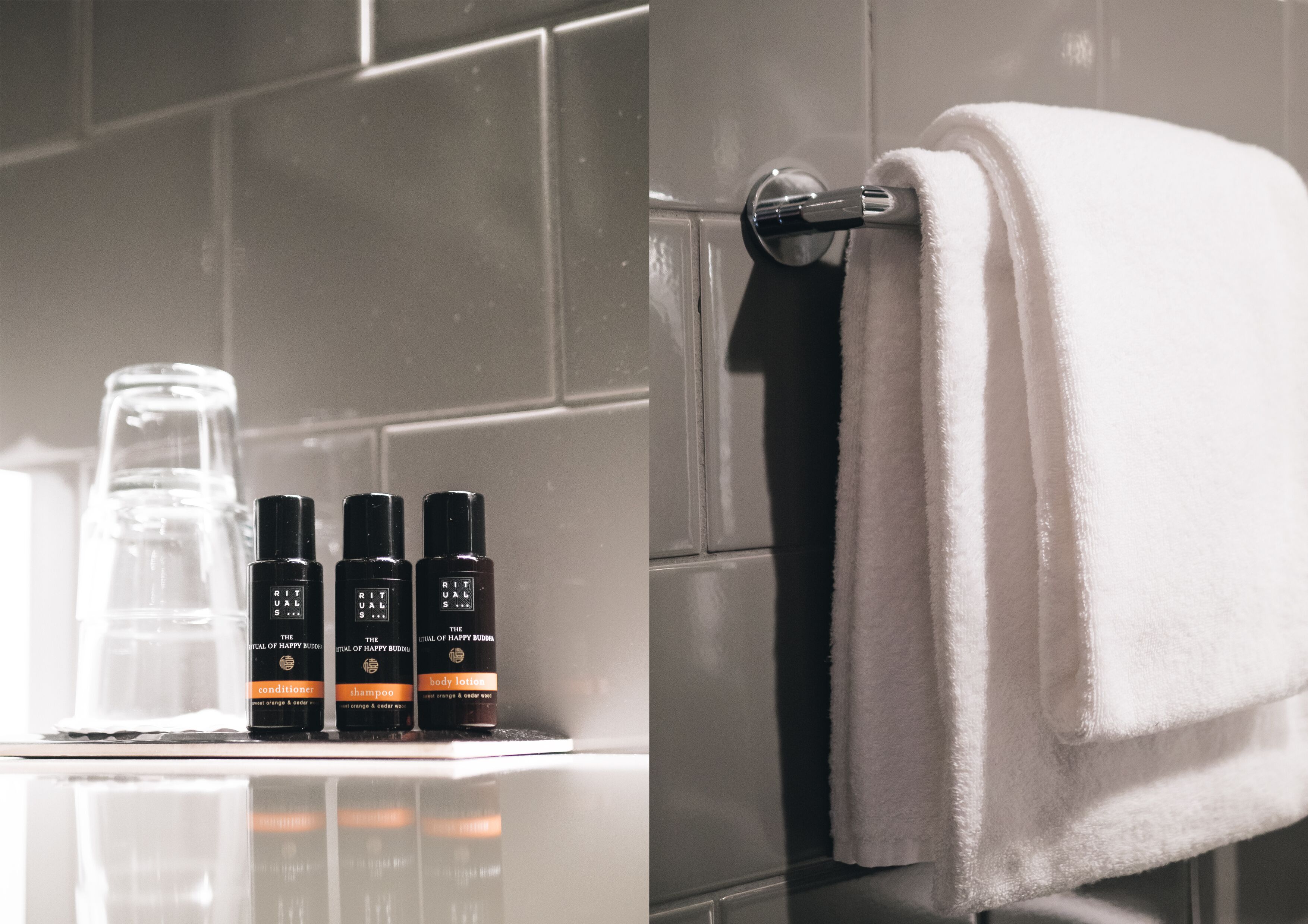 A bathroom with a white towel hanging on a rack, a glass bottle, and three black bottles labeled 'Rituals' with different contents.