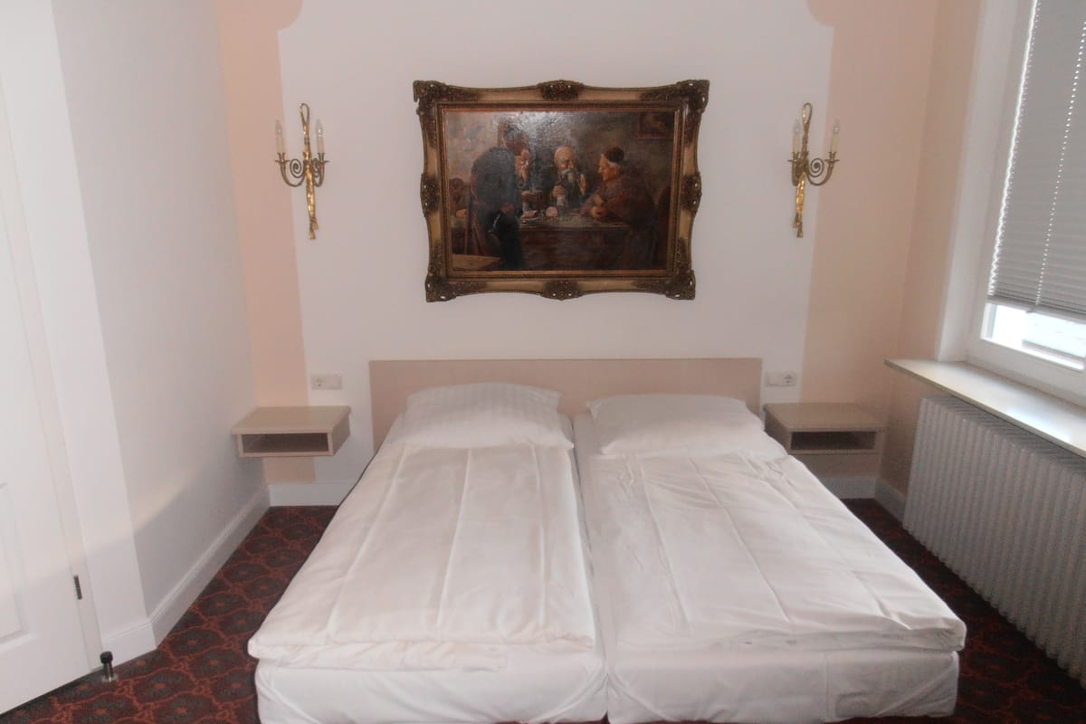 A double bed with white linens, a painting above it, and a window on the right.