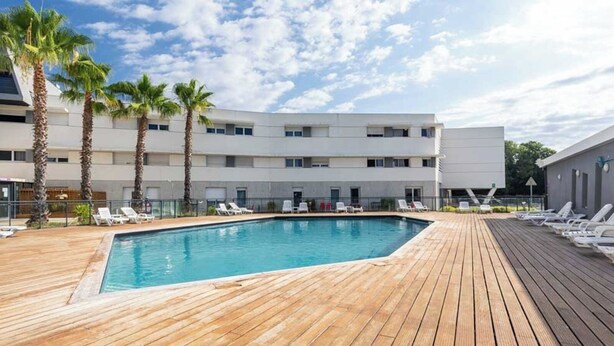 A swimming pool with wooden decking, palm trees, and a modern building with multiple windows.