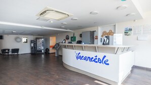 A reception desk with 'Vacanzele' signage, a vending machine, a seating area with chairs, and a clock on the wall.