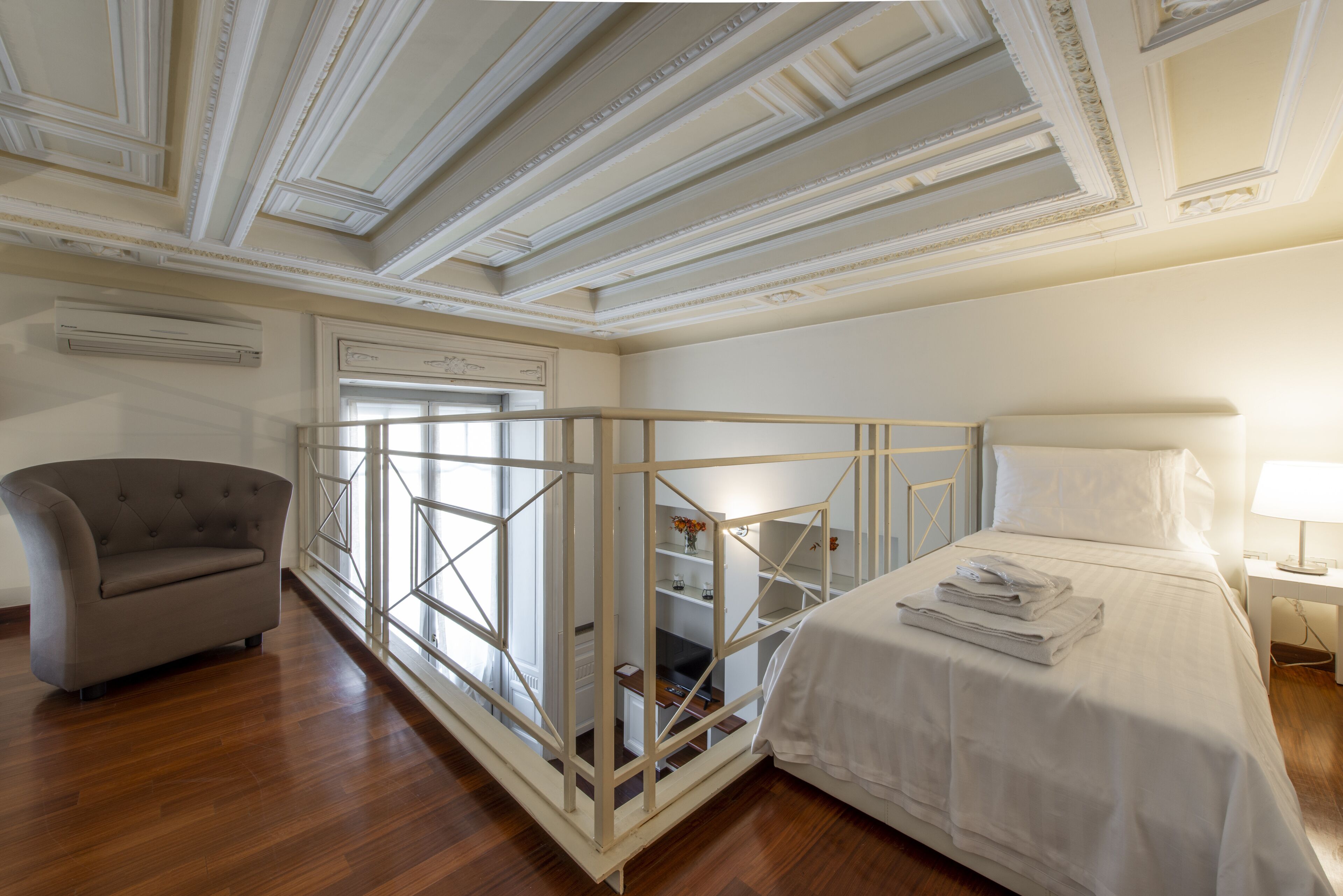 Une chambre d’hôtel avec un plancher en bois, un lit avec du linge de lit blanc, un fauteuil gris et une balustrade décorative blanche.
