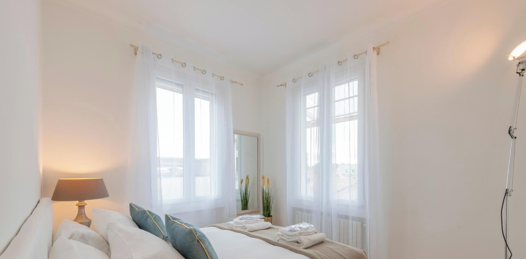 A bedroom with a bed, a bedside lamp, and a window with sheer curtains.