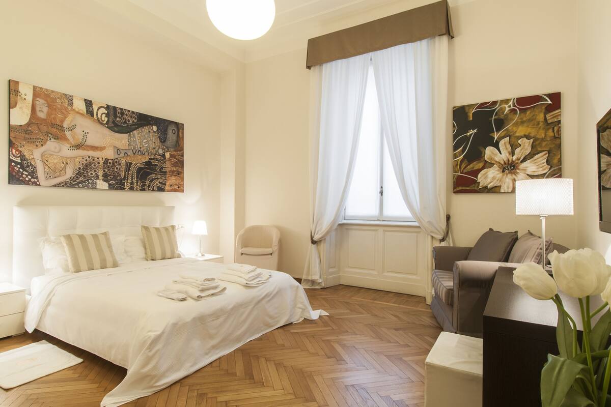 A hotel room with a bed, a chair, a sofa, a painting, and a window with curtains.