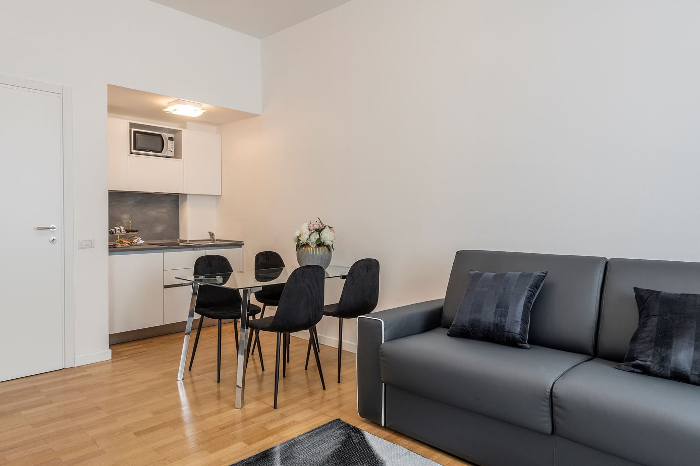 A modern living room with a grey sofa and black chairs, a kitchen with white cabinets and a microwave, and a wooden floor.