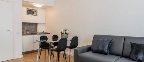A modern living room with a grey sofa and black chairs, a kitchen with white cabinets and a microwave, and a wooden floor.