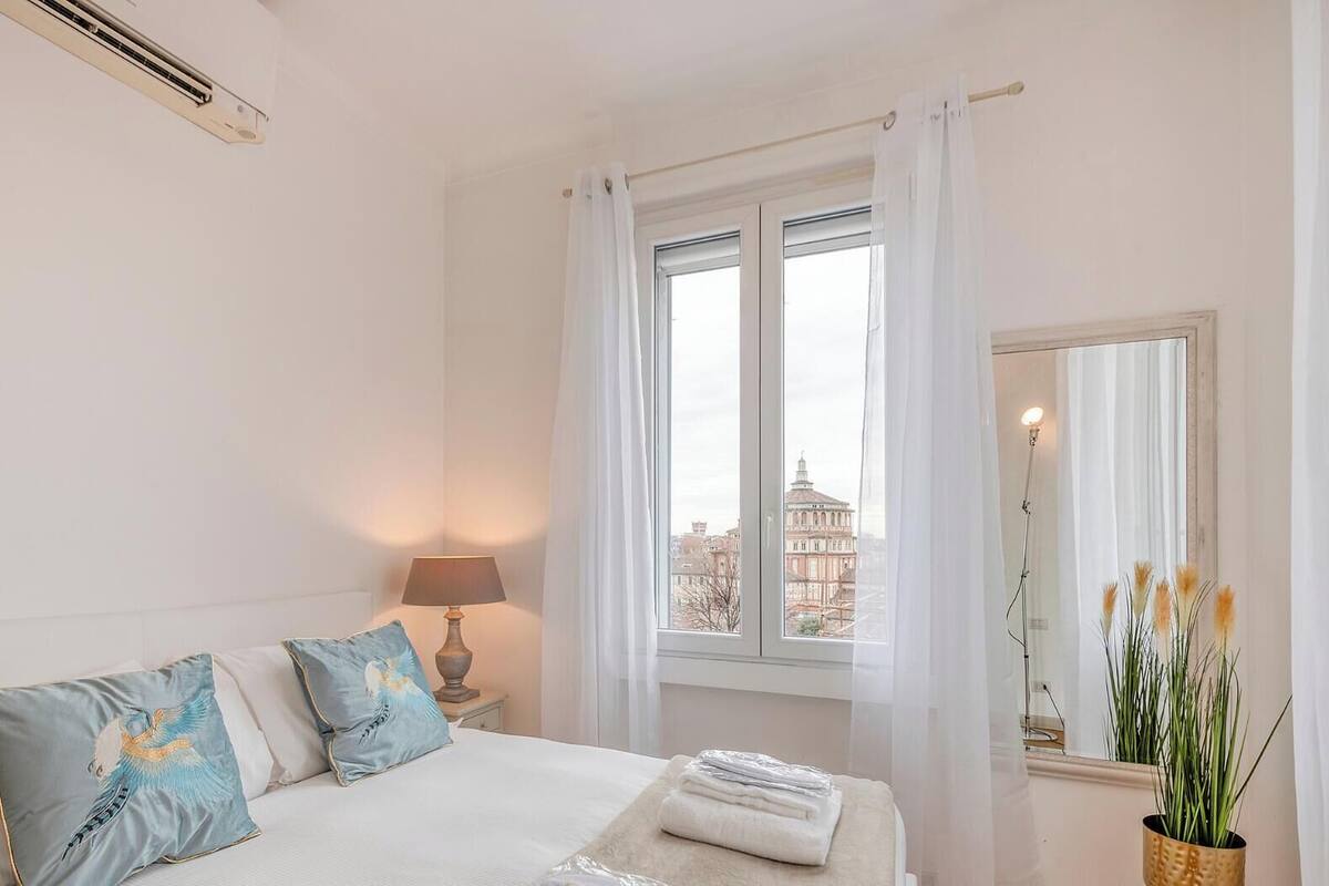 A bedroom with a bed, a bedside lamp, a window with curtains, and a view of buildings outside.