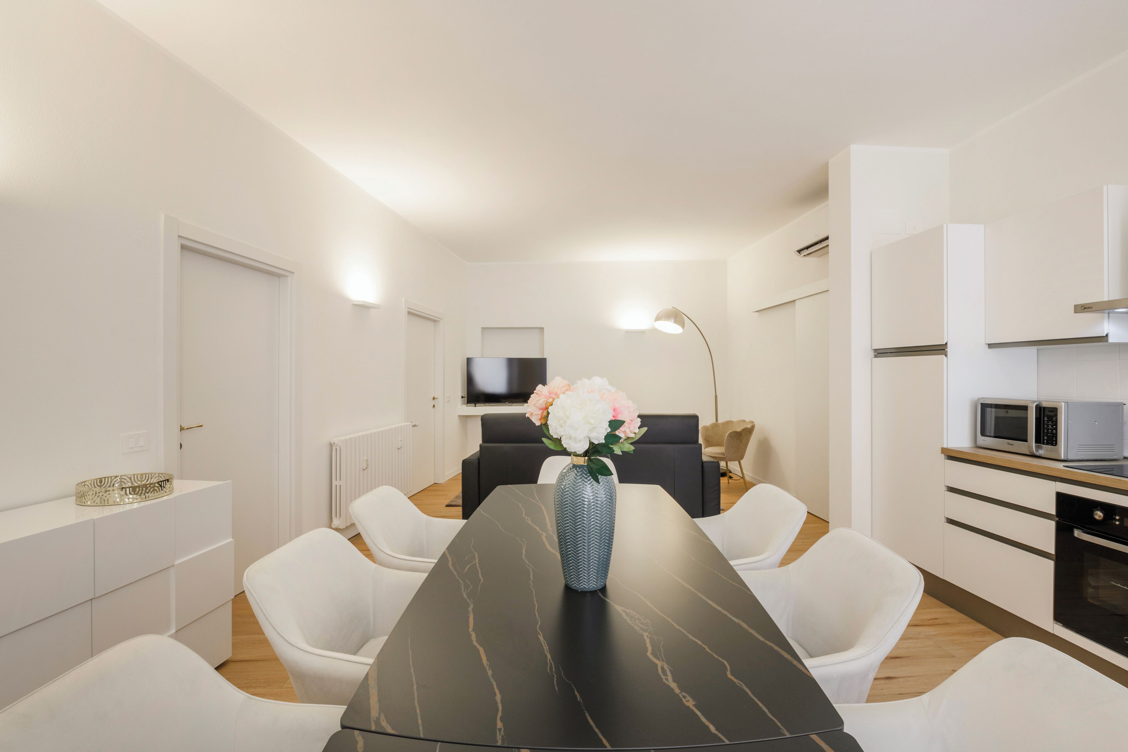 A modern kitchen with a built-in microwave and oven, a dining area with a marble table and white chairs, and a living area with a sofa and a floor lamp.