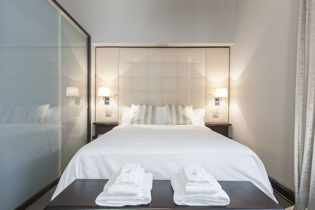 A hotel room with a large bed, two bedside lamps, a glass partition, and two folded towels on the nightstand.