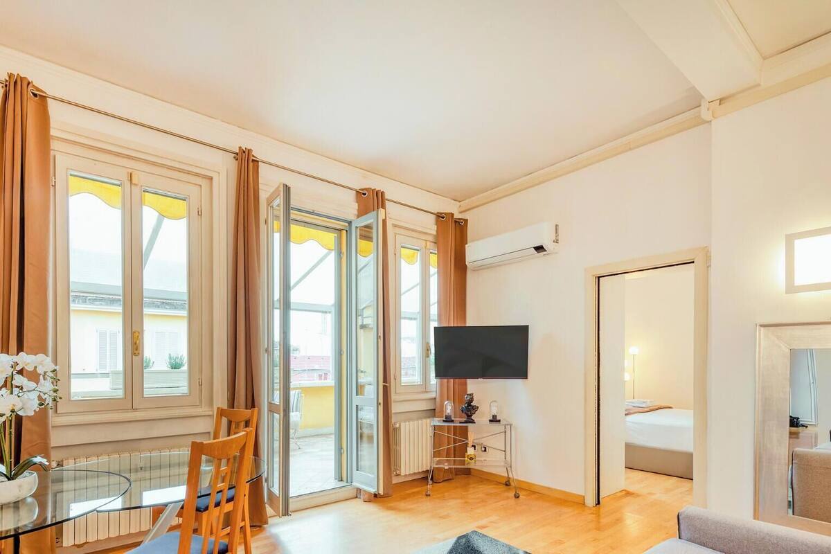 A hotel room with a dining table, chairs, a TV, a mirror, and large windows with curtains.