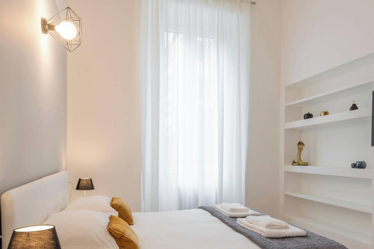 A modern hotel room with a bed, bedside lamps, a TV on a wall, and a window with sheer curtains.