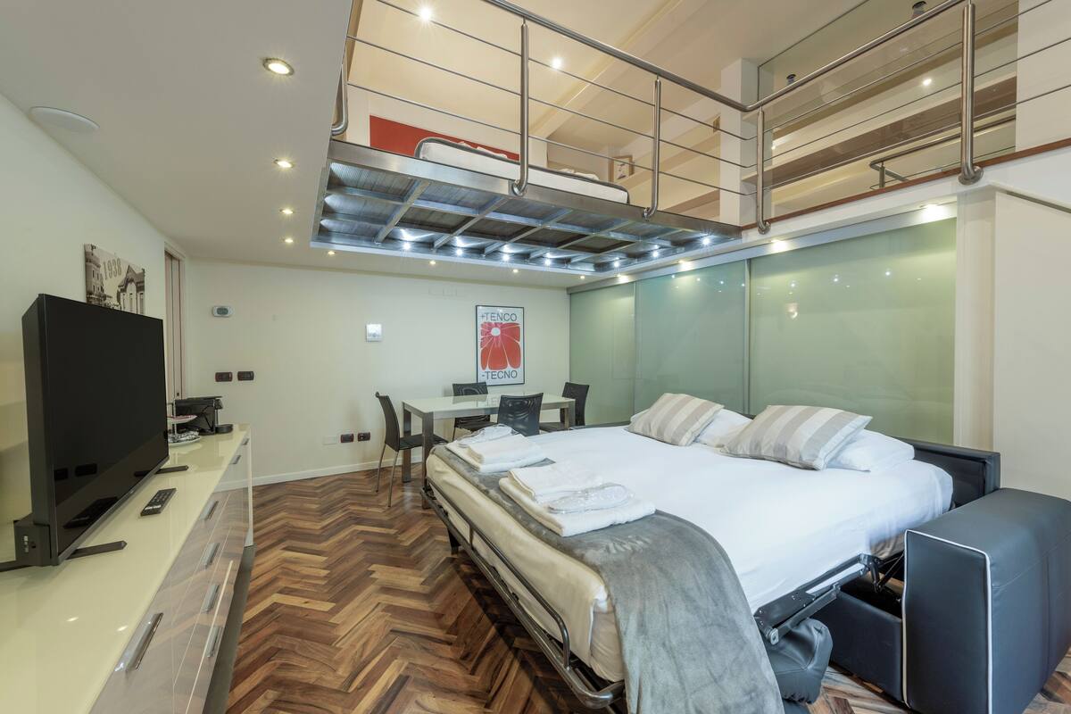 A modern hotel room with a bunk bed, a desk with chairs, a flat-screen TV, and a large bed with pillows.