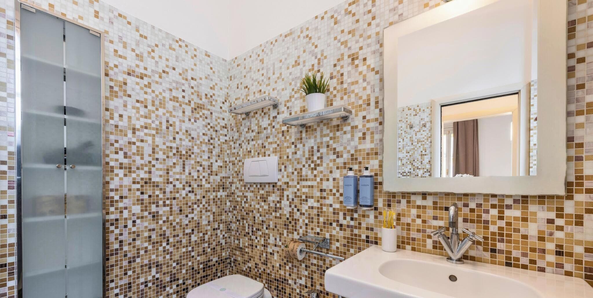 A bathroom with mosaic tile walls, a toilet, a sink, a mirror, and a shower enclosure.