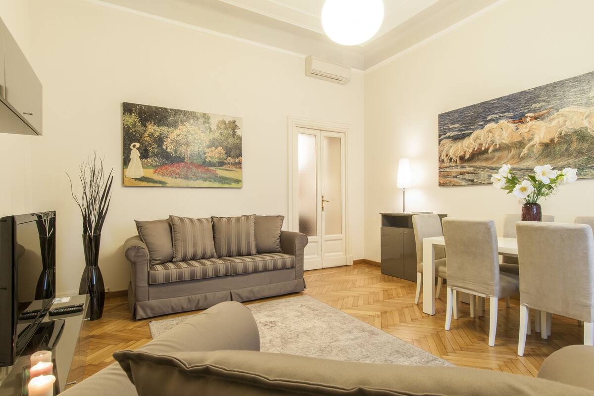 A living room with a grey sofa, a dining table with chairs, a TV, and two paintings on the walls.