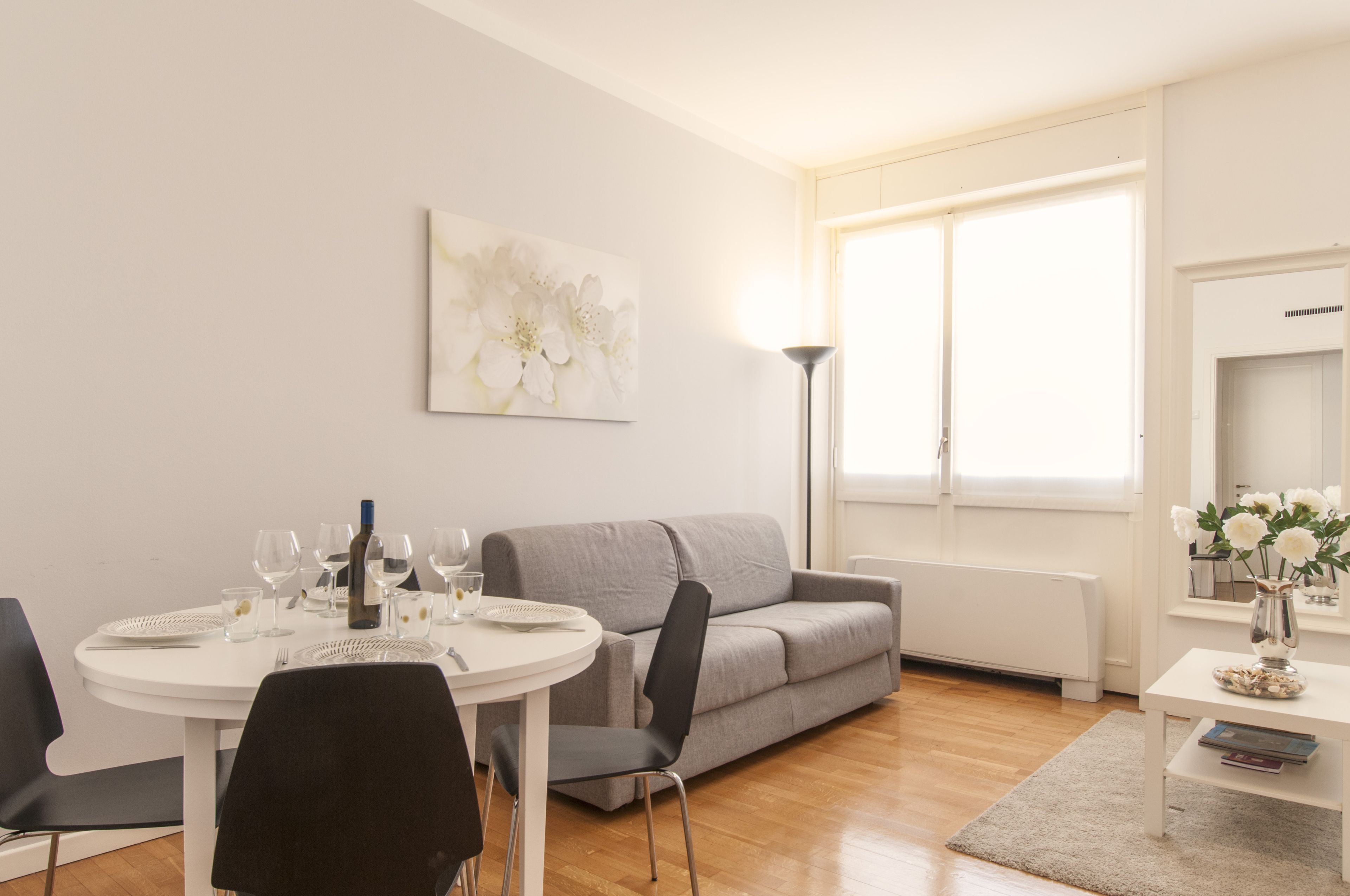 A modern living room with a dining table set for four, a grey sofa, and a large window with natural light.
