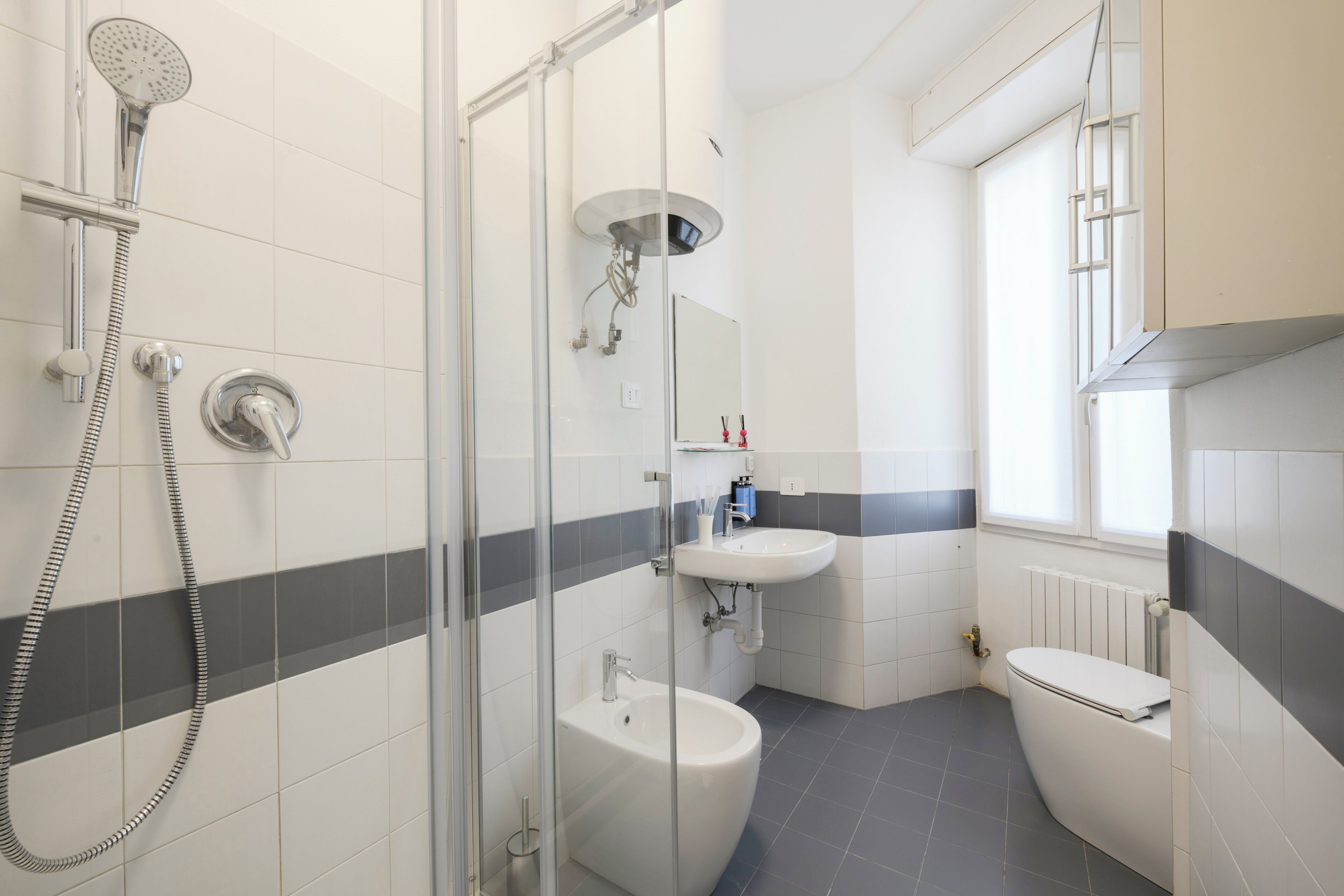 Un baño moderno con cabina de ducha de vidrio, un lavamanos blanco, un inodoro y una ventana que deja entrar luz natural.