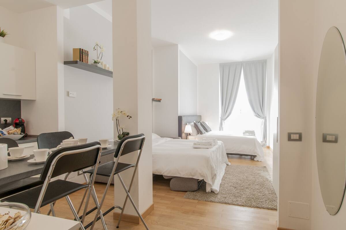 A modern hotel room with a dining area, a bed with a white duvet, a bedside table with a lamp, and a large mirror.