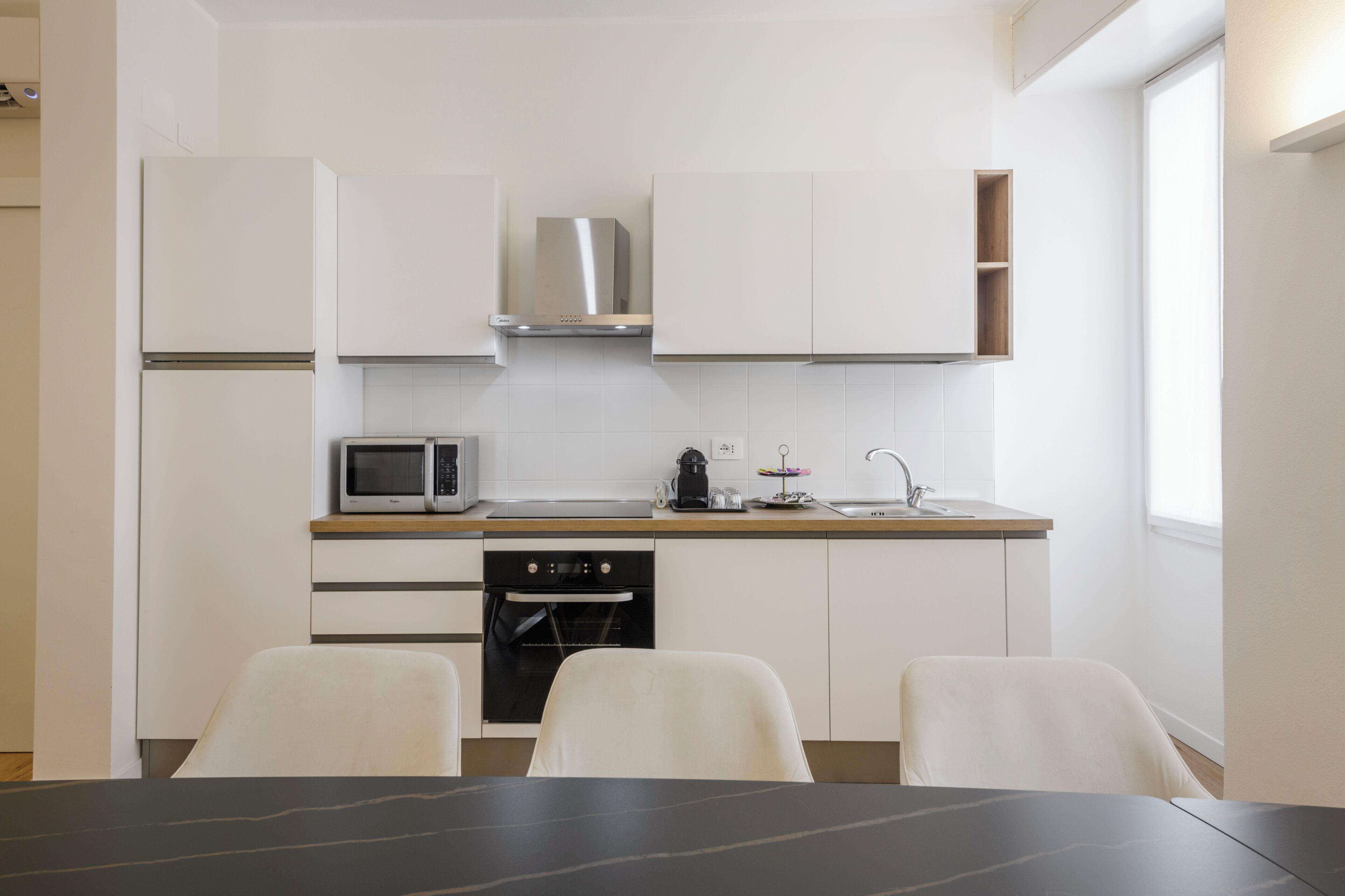 A modern kitchen with white cabinetry, a stainless steel range hood, a microwave, a built-in oven, and a sink. There are two white chairs at a table.