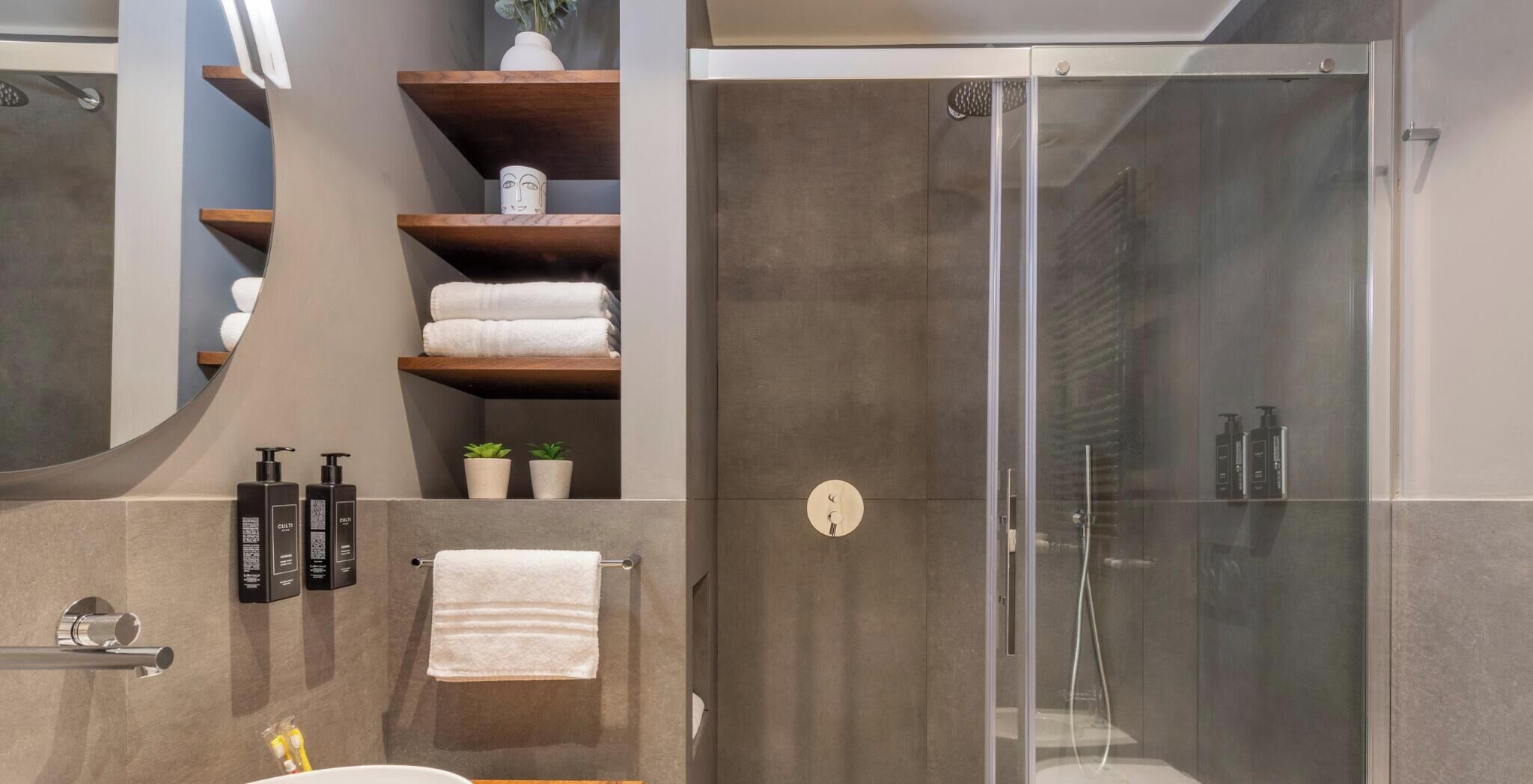 A modern bathroom with a glass-enclosed shower, wooden shelves with towels, and toiletries.