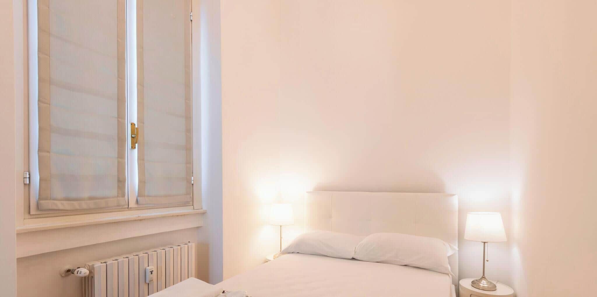 Una camera da letto con un letto, due lampade da comodino, una finestra con tende e un termosifone.