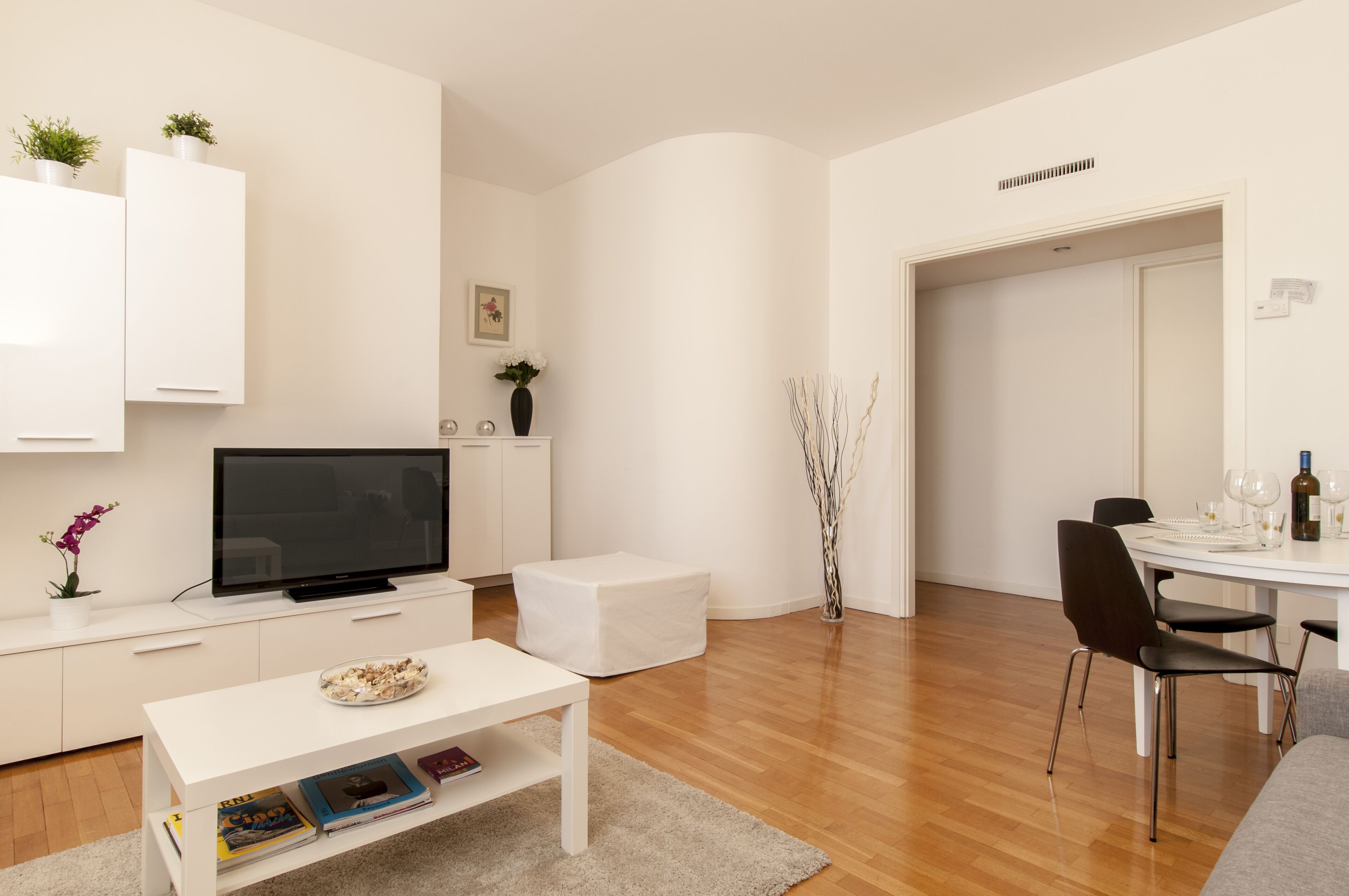 A modern living room with a flat-screen TV, white furniture, a dining area with a table and chairs, and a hallway leading to another room.