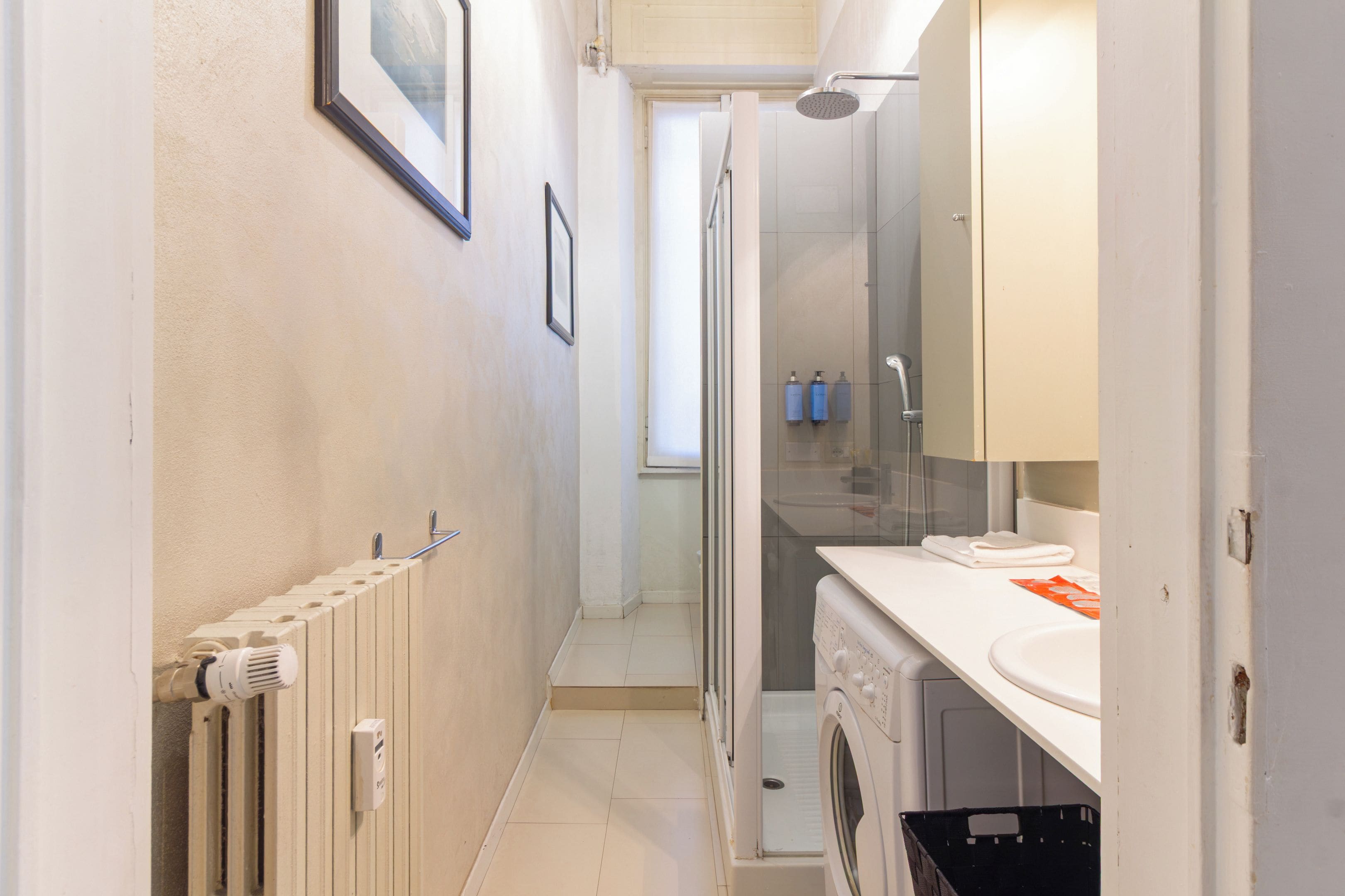 A narrow hallway leading to a bathroom with a shower, a radiator, and a washing machine.