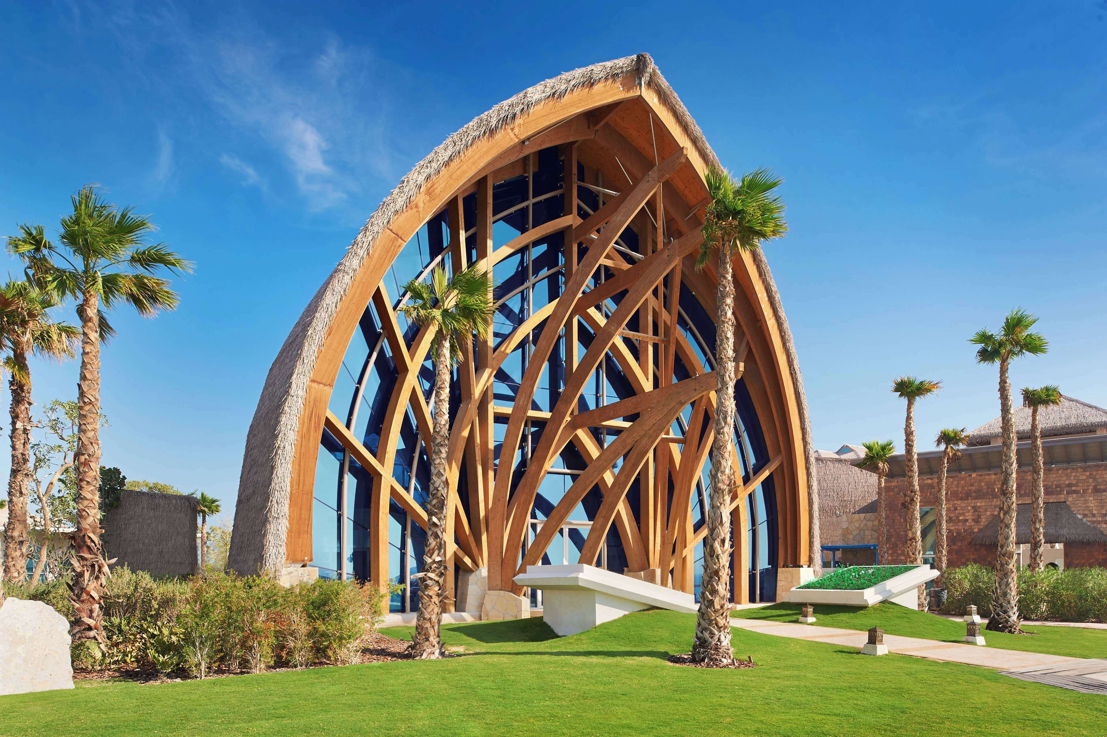 Un edificio moderno con una estructura arqueada única y vigas de madera, rodeado de palmeras y un césped bien cuidado.