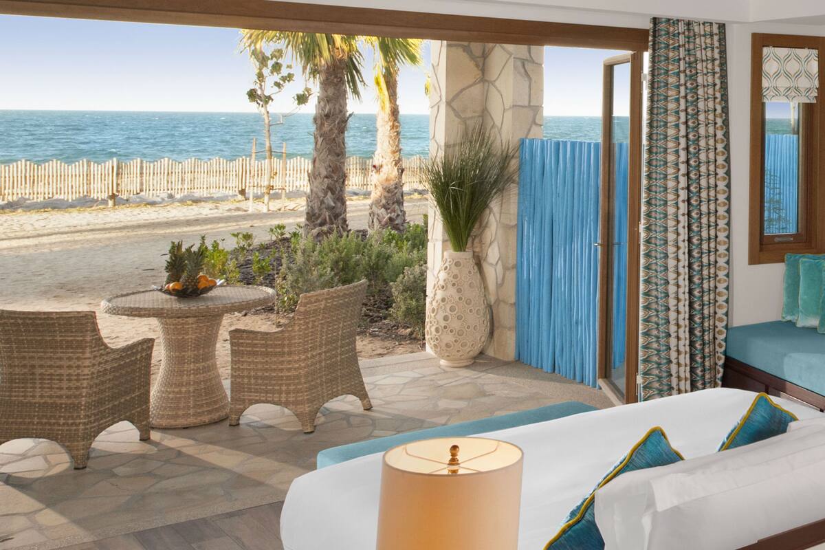 Een slaapkamer met uitzicht op het strand, met rieten meubels, een groot raam met behang en een bed met blauwe kussens.