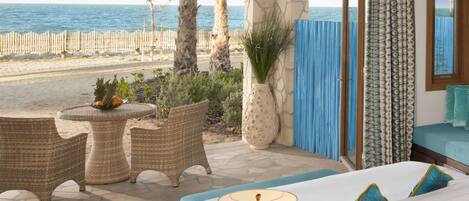 Un dormitorio con vistas a la playa, muebles de mimbre, un ventanal con cortinas estampadas y una cama con almohadas azules.