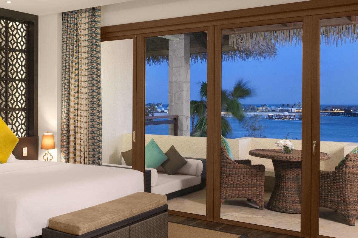 Een hotelkamer met een groot bed, een balkon met rieten meubilair en uitzicht op zee en gebouwen.