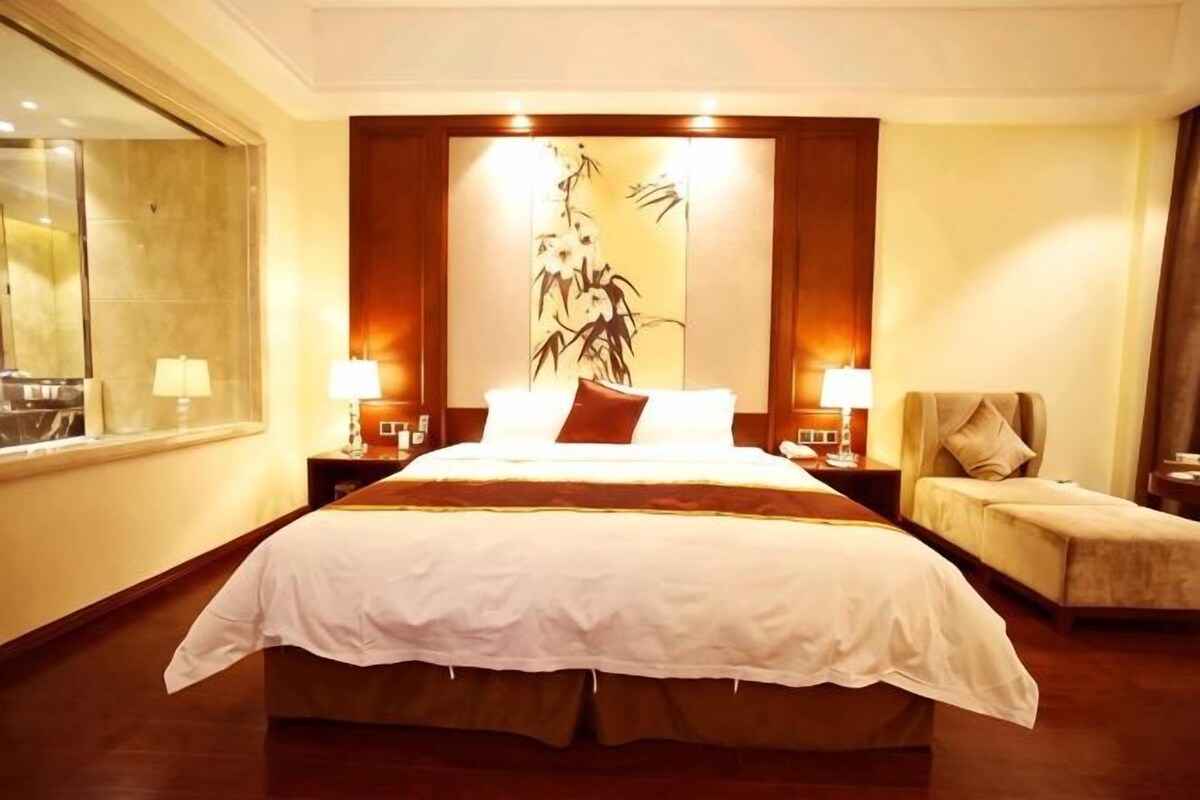 A hotel room with a large bed, two bedside tables with lamps, a chair, and a large window.