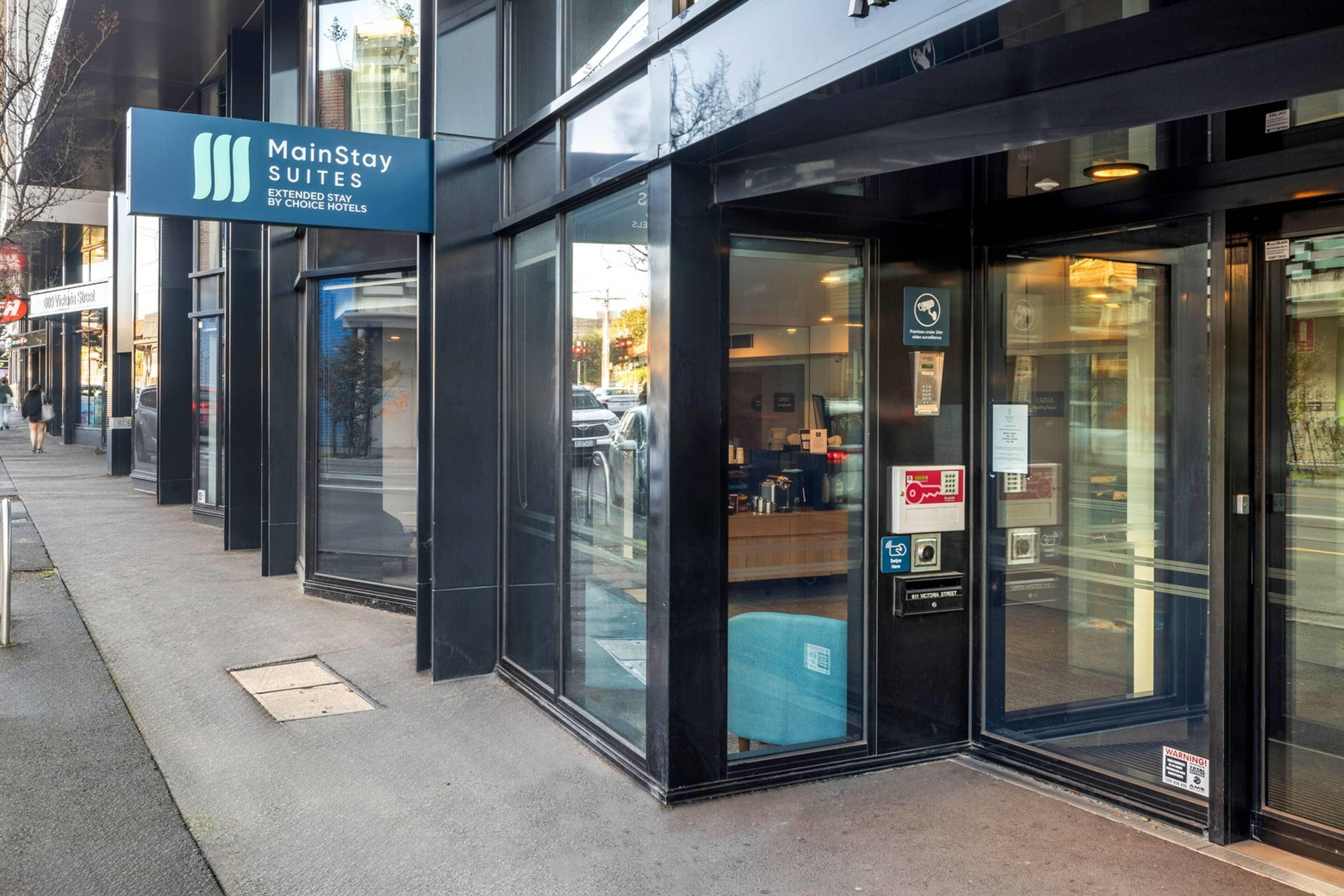 A MainStay Suites hotel entrance with glass doors, a signboard, and a security camera.