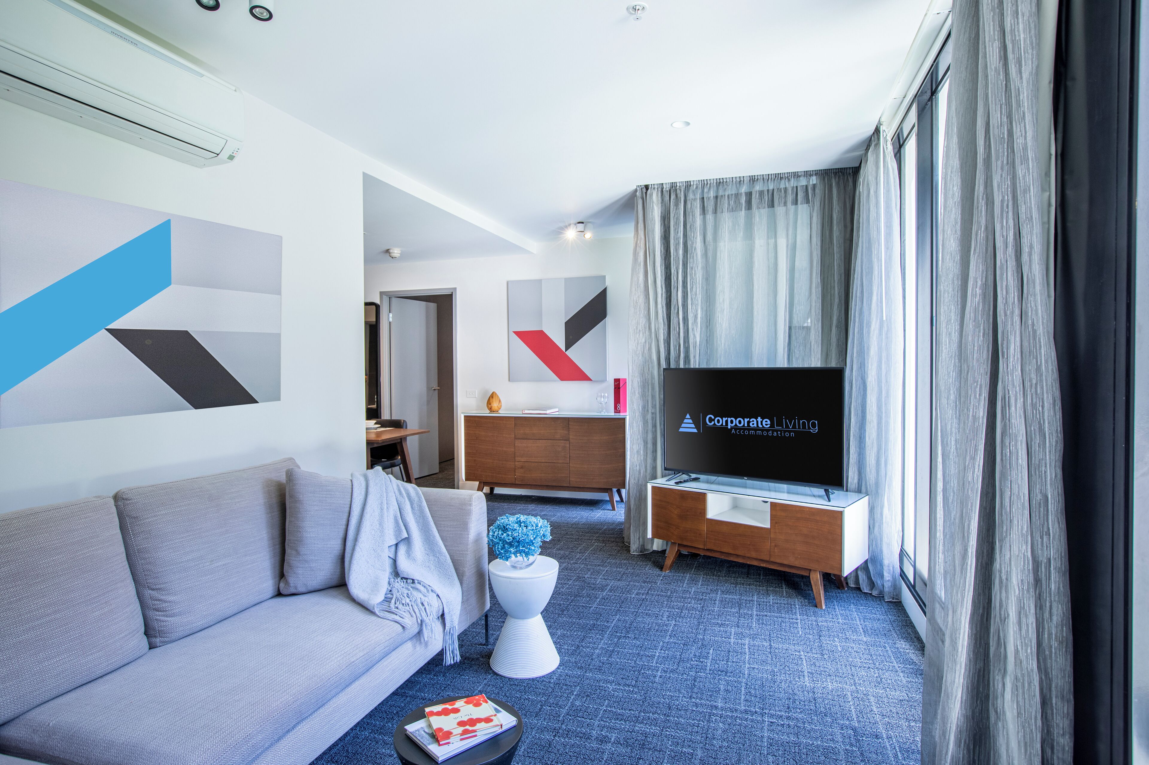 A modern hotel room with a grey sofa, a white side table with a blue vase, a wooden dresser, and a flat-screen TV displaying "Corporate Living".