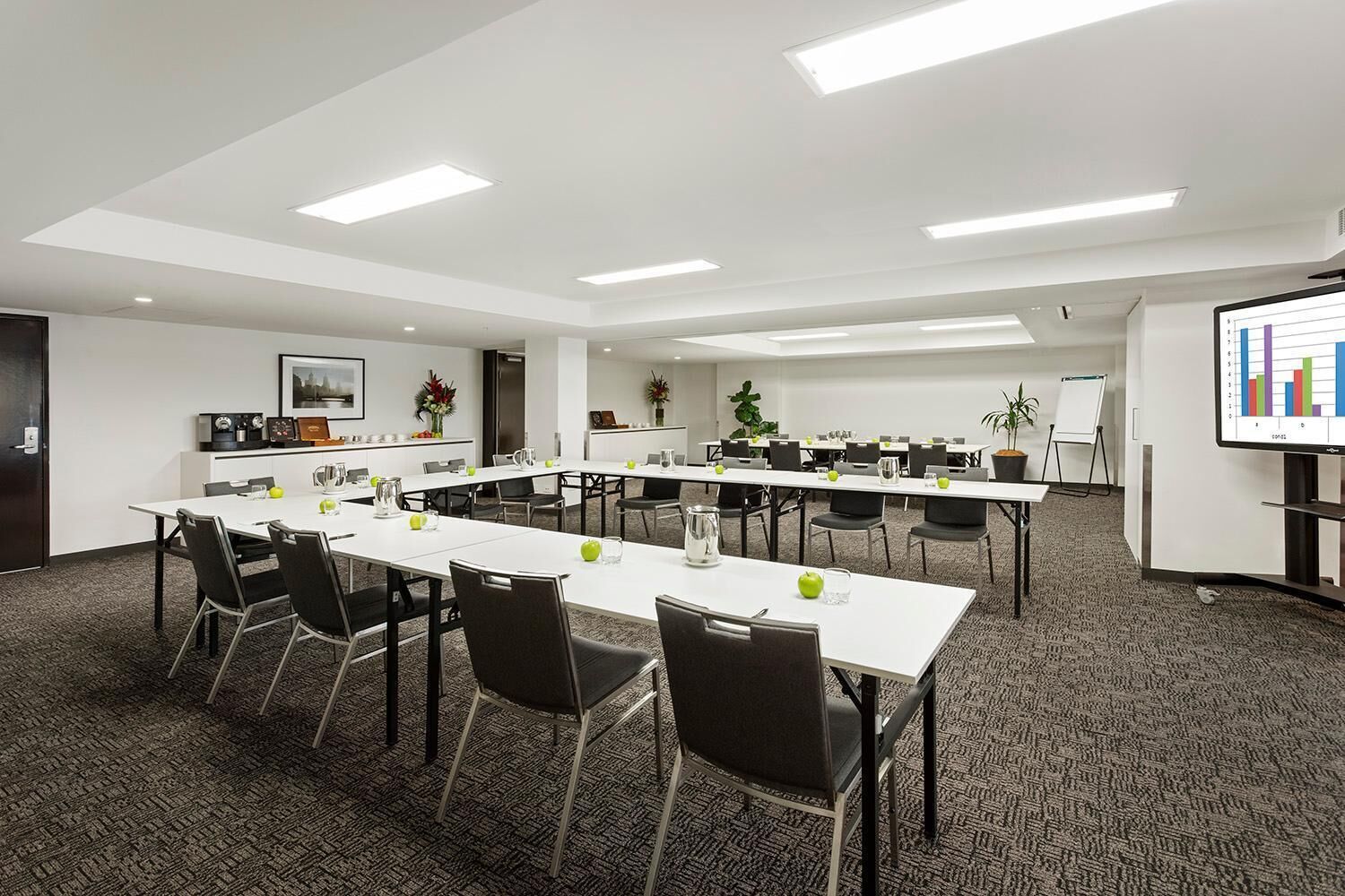 Un salón de conferencias con mesas blancas, sillas negras y una pantalla proyectando un gráfico de barras.