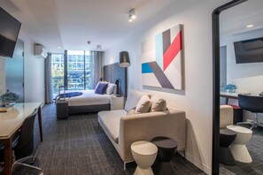 Hotellihuone, jossa on sänky, työpöytä, sohva, televisio ja suuri ikkuna.