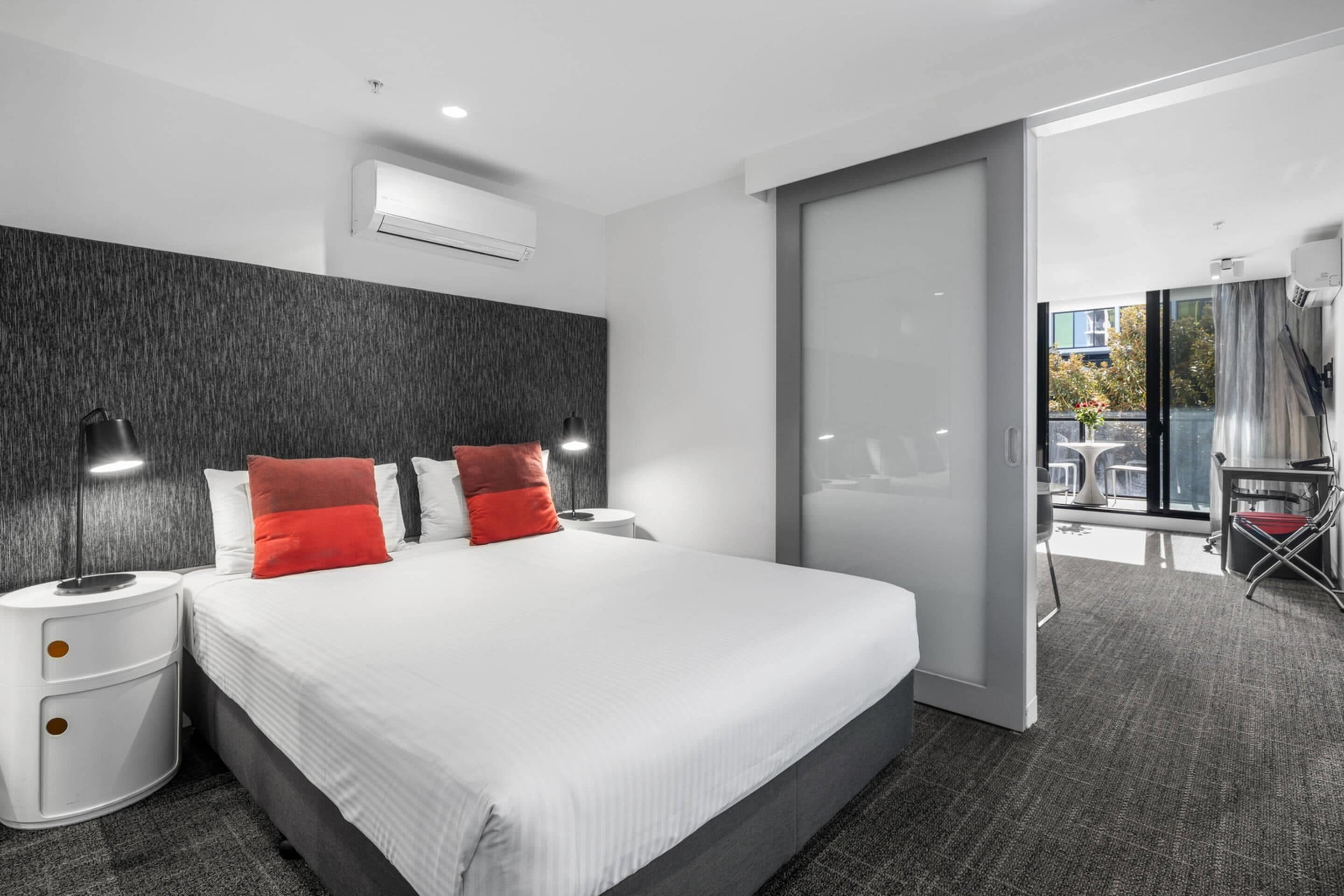 A modern hotel room with a large bed, two bedside tables, a wardrobe, and a sliding glass door leading to a balcony with outdoor furniture.