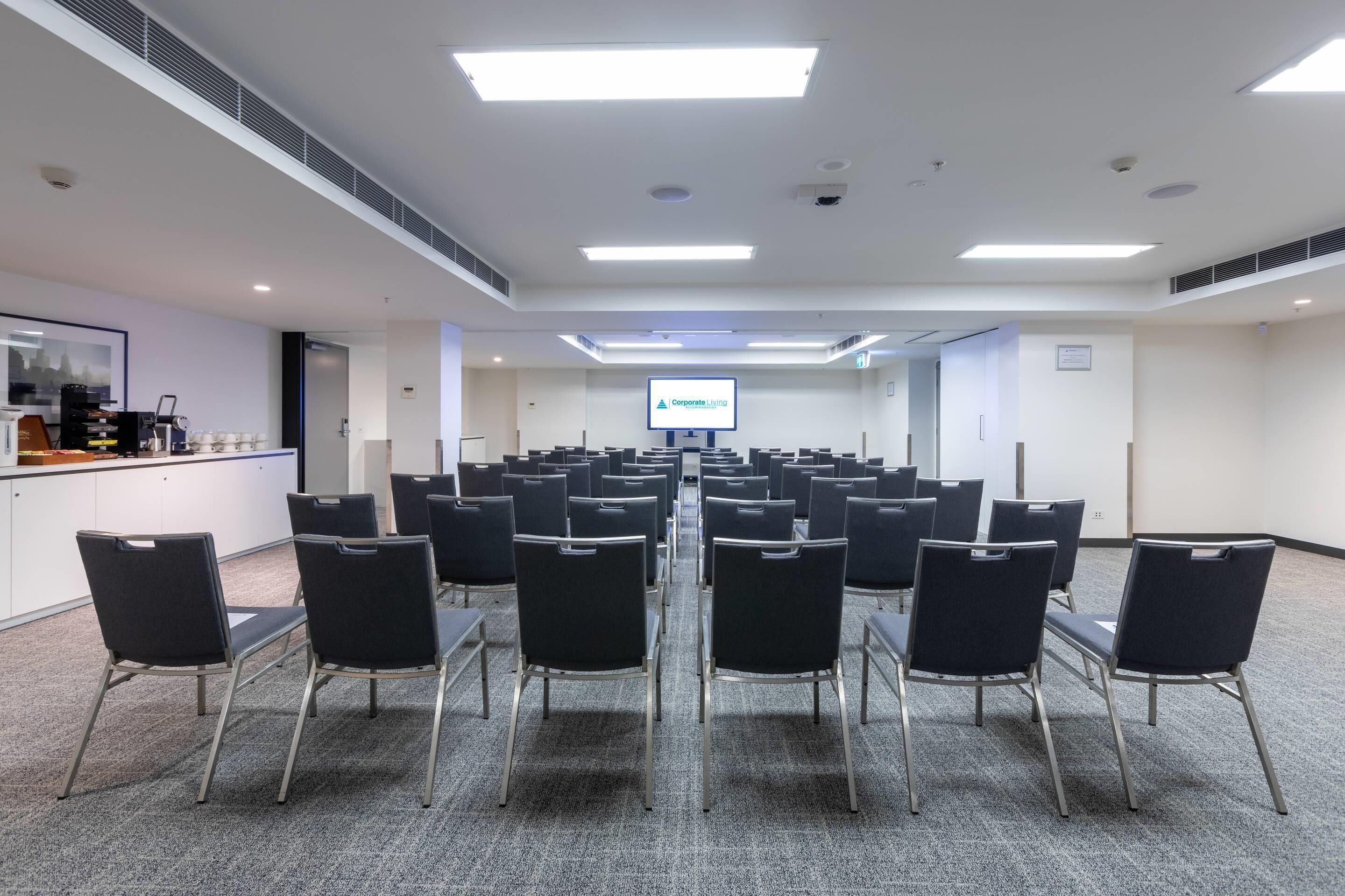 Une salle de conférence avec des rangées de chaises, un coin café et un écran de présentation.