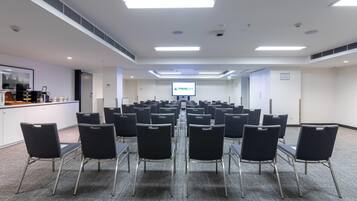 Un salón de conferencias con filas de sillas, una estación de café y una pantalla para presentaciones.