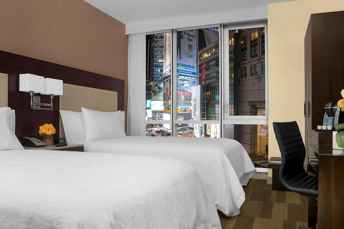 Habitación de hotel con dos camas, un escritorio, una silla y vistas a un paisaje urbano a través de amplias ventanas.