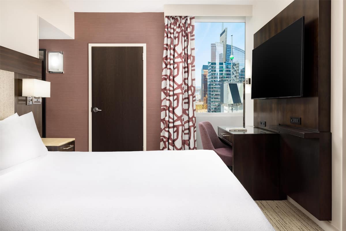 Una habitación de hotel con cama, escritorio, televisor, ventana y vista a la ciudad, además de una puerta.