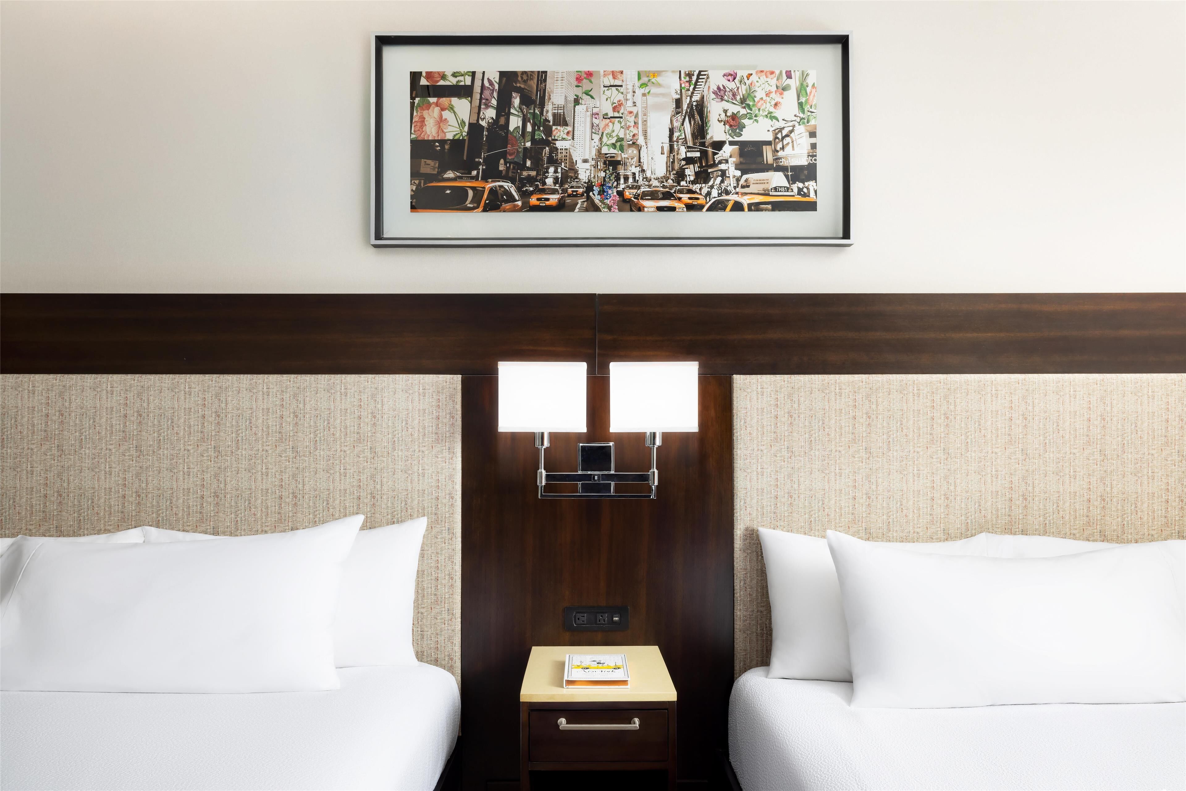 A hotel room with two beds, a framed cityscape picture, a nightstand with a book, and a control panel.