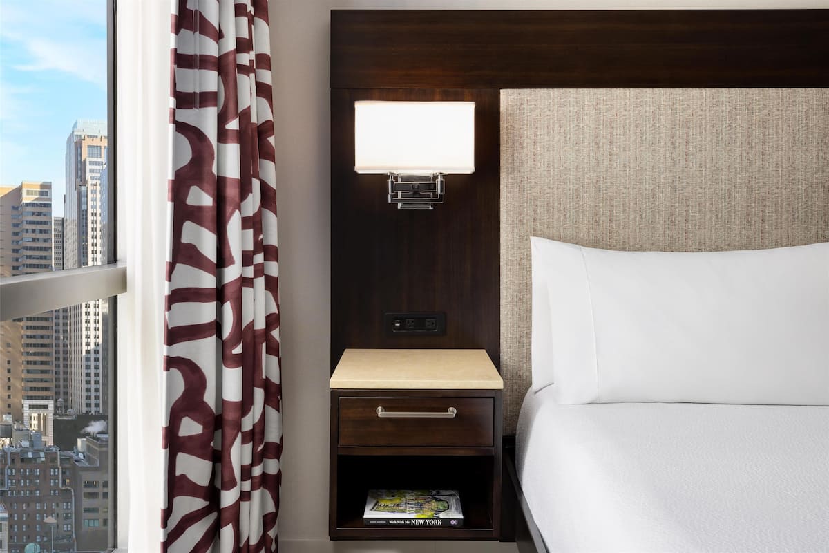 Habitación de hotel con cama, mesita de noche con lámpara, cajón y estante con una revista.