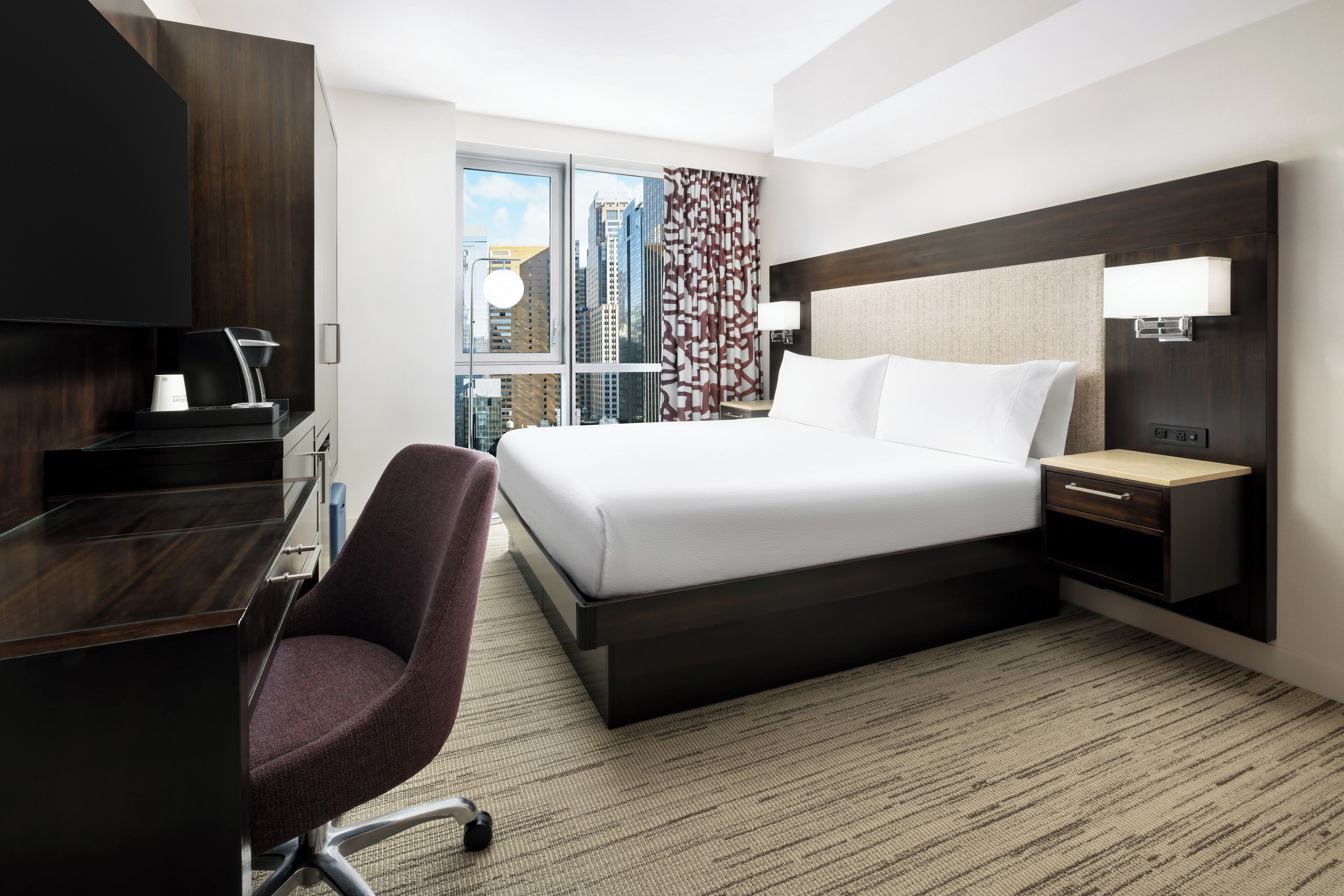 A hotel room with a large bed, a desk with a chair, a nightstand, and a view of the city through the window.