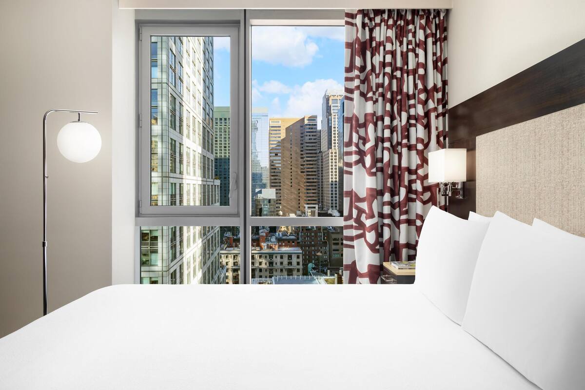 Una habitación de hotel moderna con un ventanal grande, una cama con ropa blanca, una lámpara de pie y una cortina estampada.