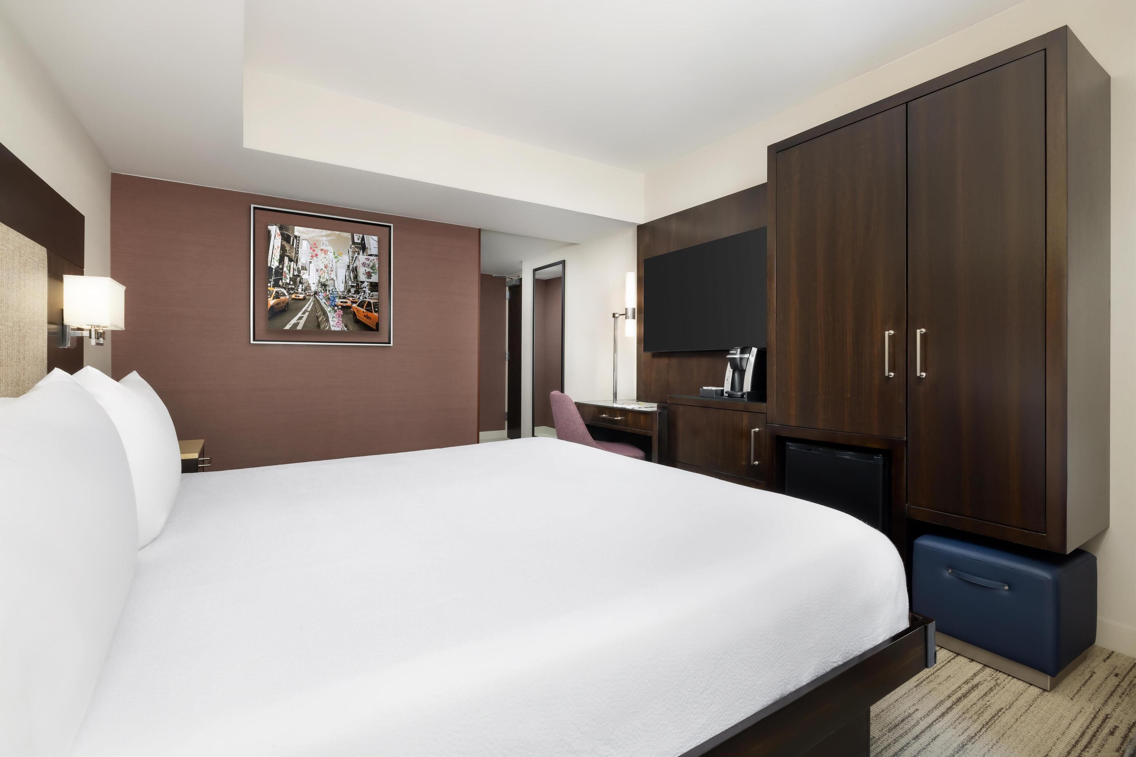 A hotel room with a large bed, a desk with a chair, a wardrobe, and a flat-screen TV mounted on the wall.