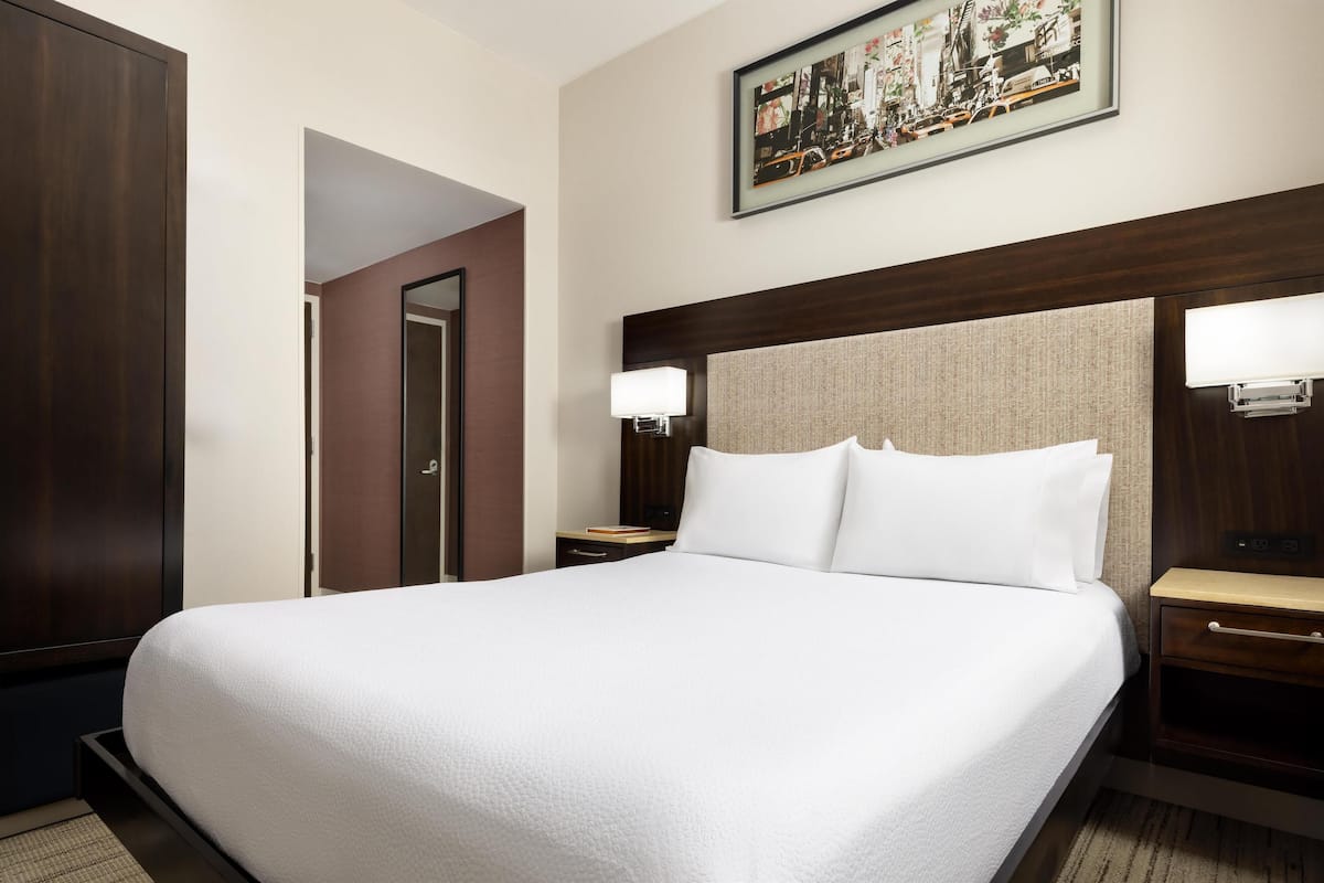 Un cuarto de hotel con una cama grande, dos mesitas de noche con lámparas, un cabecero de madera y un cuadro enmarcado en la pared.