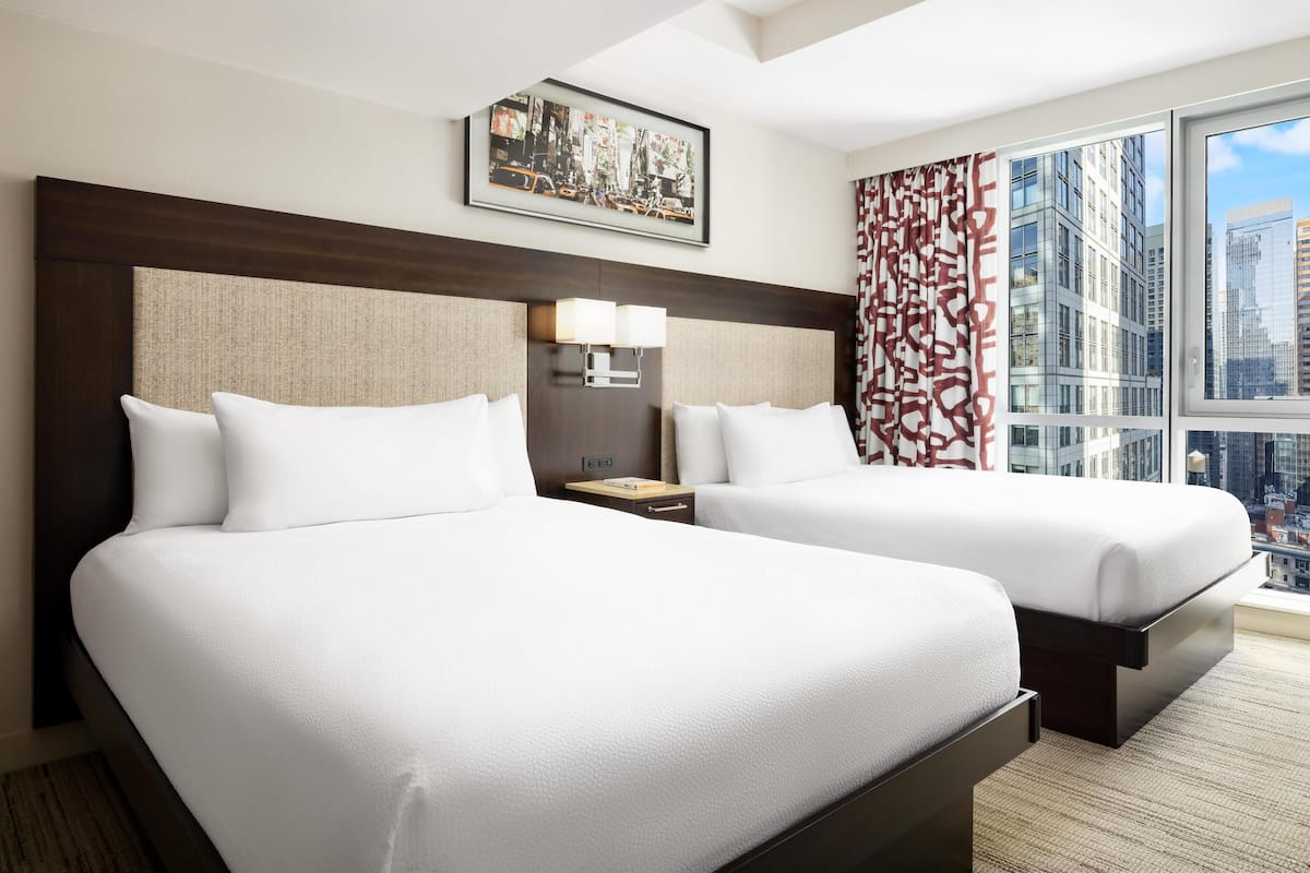 Habitación de hotel con dos camas, un ventanal con vistas a la ciudad, un cuadro en la pared y una cortina estampada.