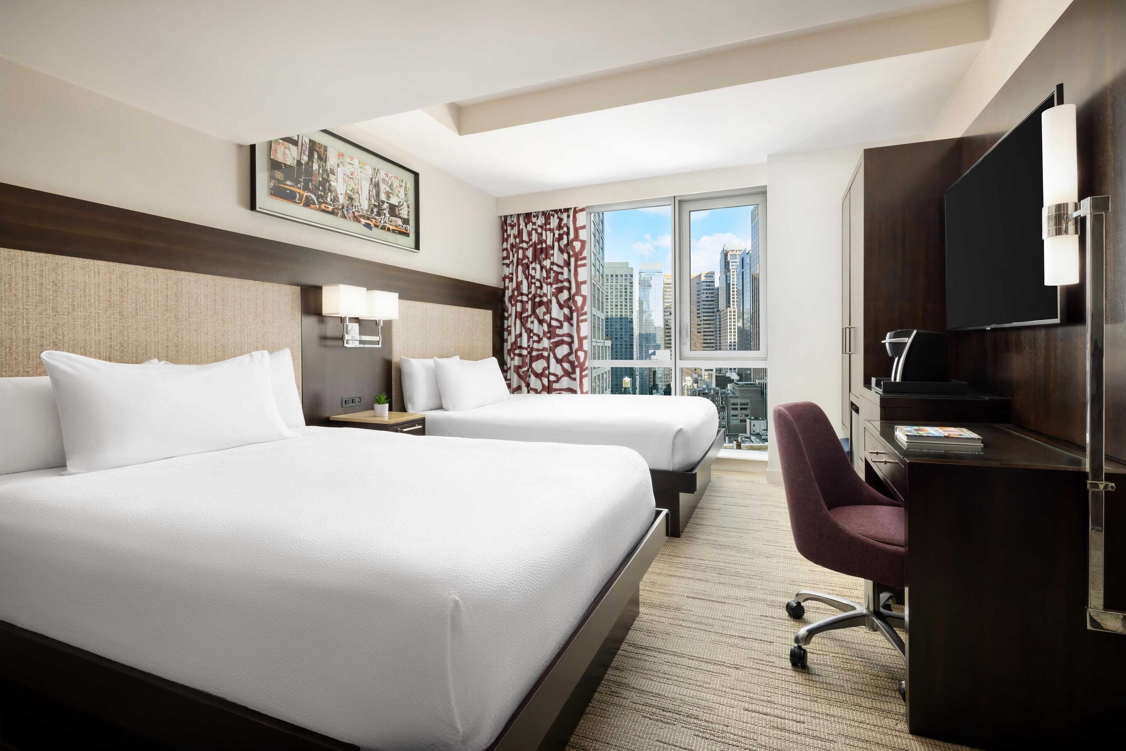 A hotel room with two beds, a desk, a chair, a TV, and a city view.