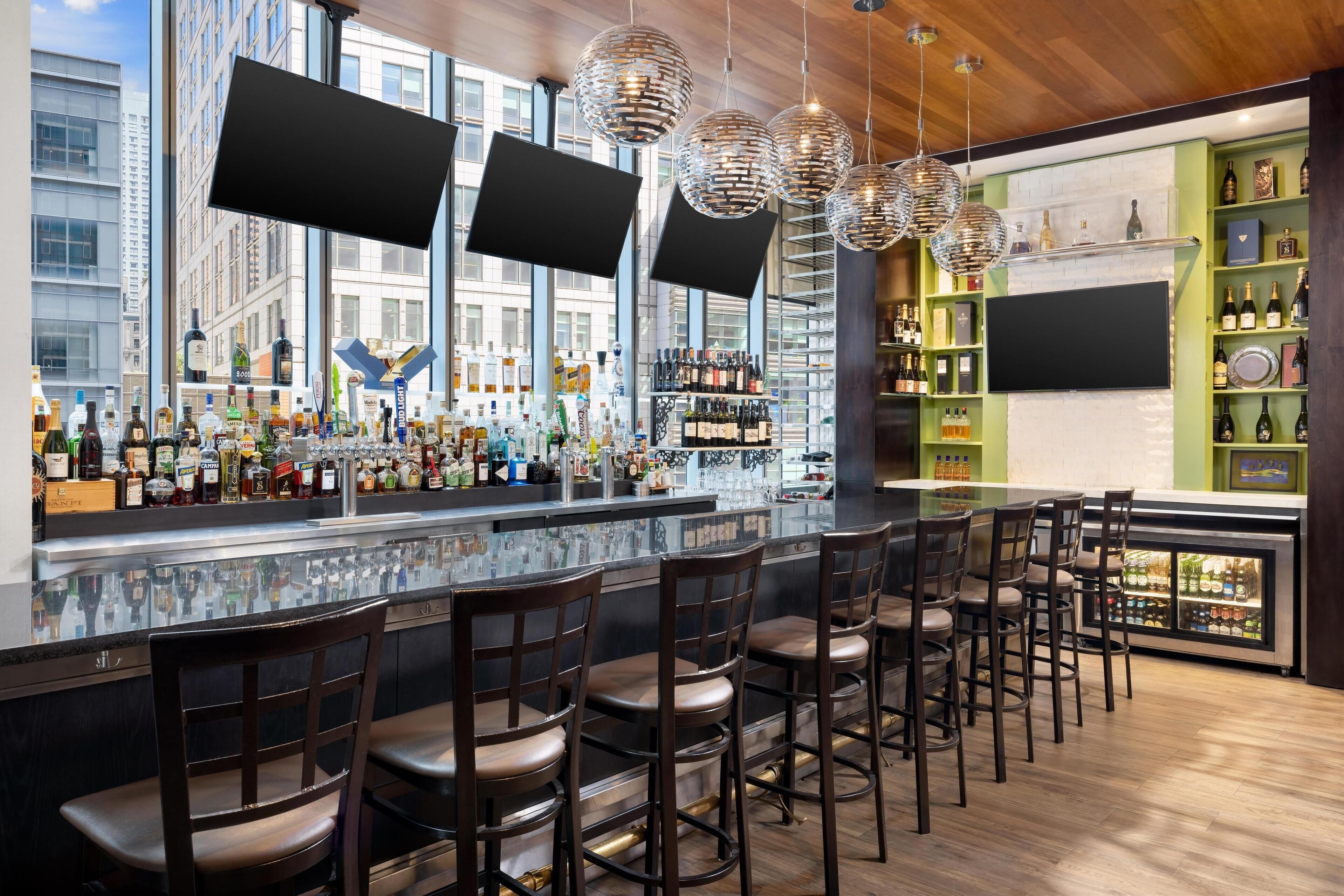 A modern bar with a long counter, multiple bar stools, and a variety of bottles and glasses on shelves.